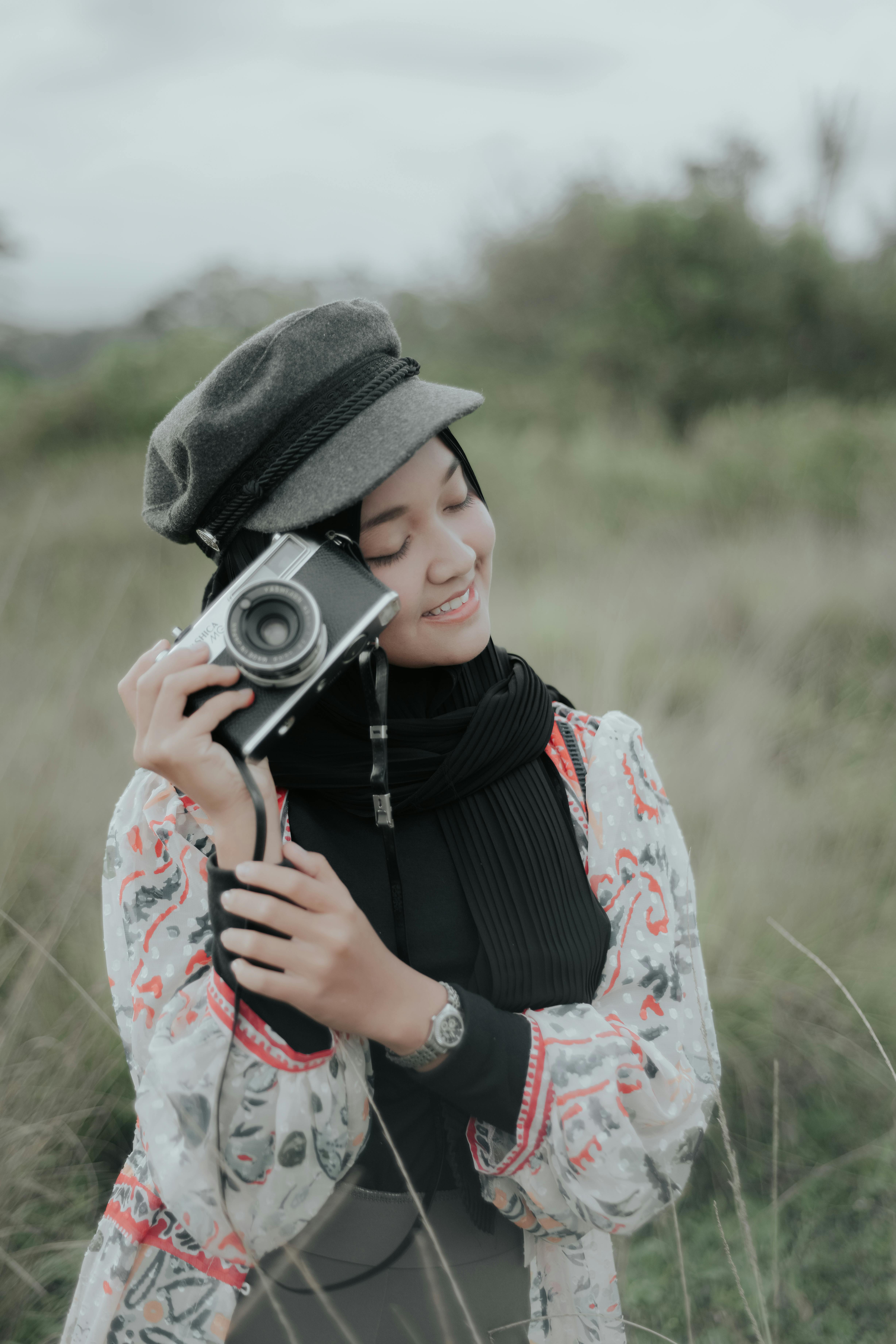 A woman wearing a hijab smiles while holding a vintage camera in a grassy field, capturing tranquil moments outdoors.