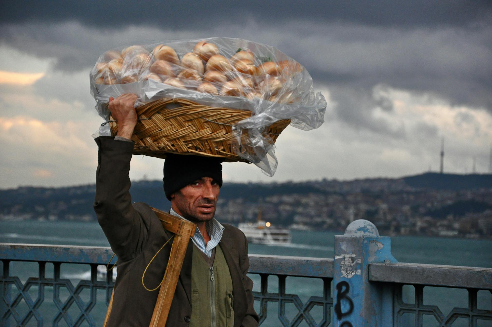 A street vendor in Istanbul carries a basket of bread on his head with a cityscape backdrop.