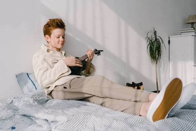 A Boy Playing Ukelele While Sitting On A Bed