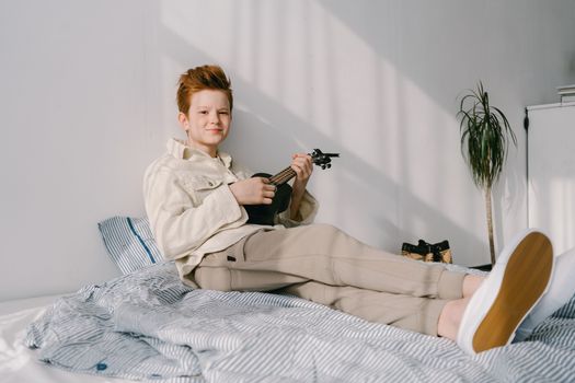 A preteen redhead boy plays a ukulele while relaxing on a bed in a sunlit room.