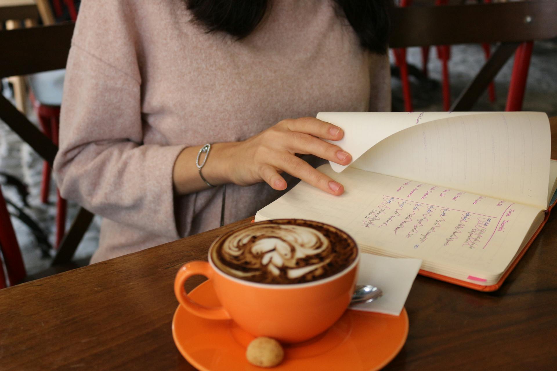 A woman flips pages in a journal beside a cappuccino on a wooden table in a cozy cafe.