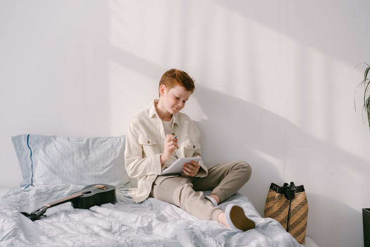 A Boy Holding A Pen And Notebook While Sitting On A Bed