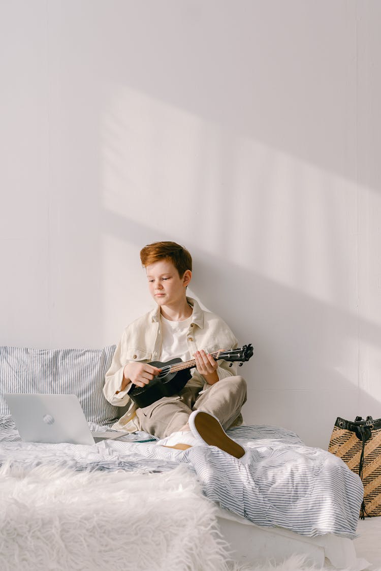 A Boy Playing Ukelele While Sitting On A Bed