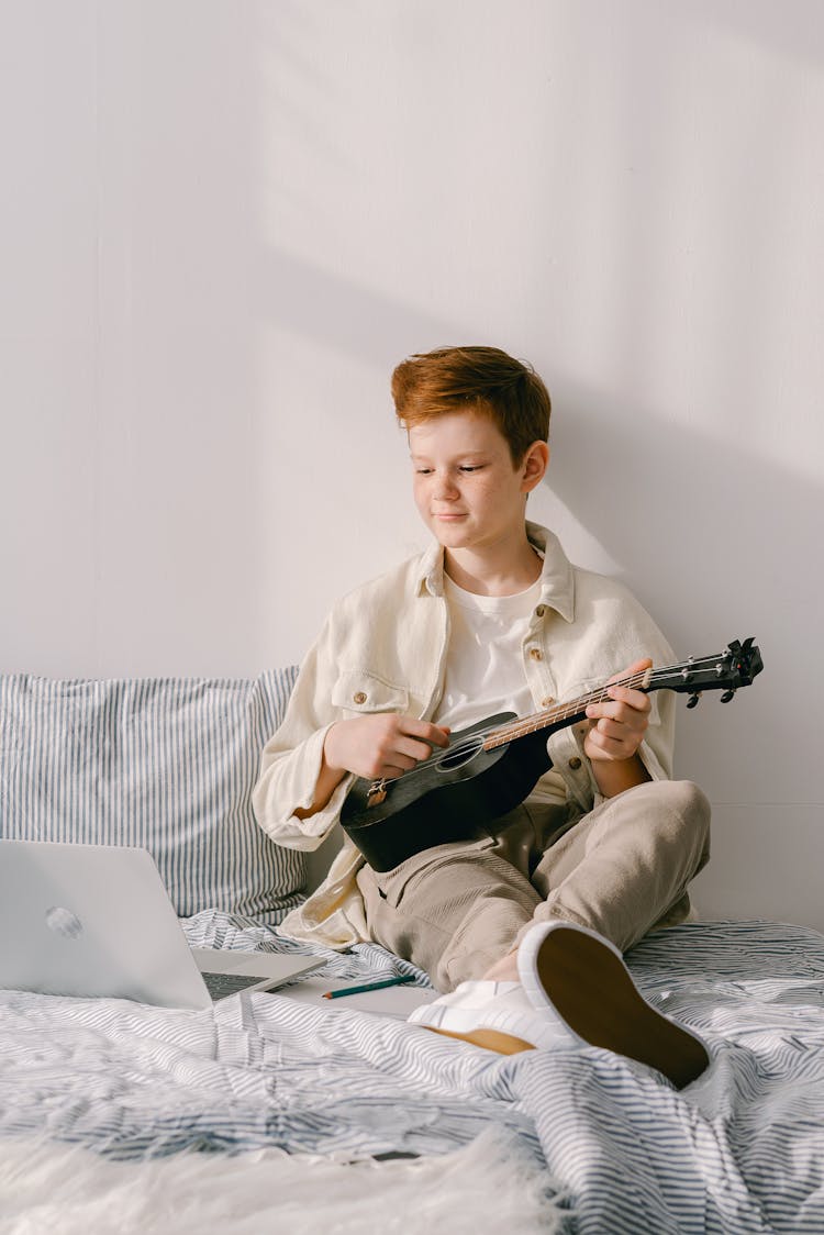 A Boy Playing Ukelele While Sitting On A Bed
