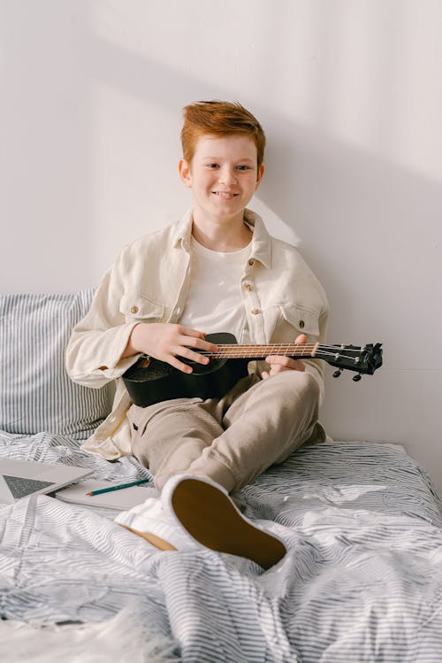 Free A Young Boy Sitting on His Bed while Holding a Ukulele Stock Photo