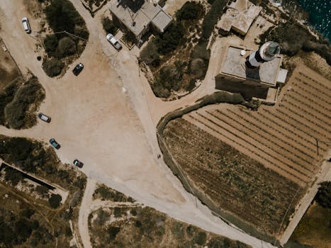 Drone photo capturing a lighthouse and surrounding fields in Marsaxlokk, Malta, with nearby roads and vehicles.