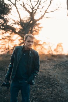 A young caucasian man in a leather jacket holding a camera at sunset in the English countryside.