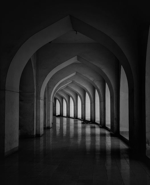 Grayscale image of an empty corridor with arched architecture and reflected shadows.