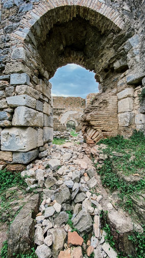 Aged stone arch construction located on grassy ground with rocks against ruins of ancient buildings on summed day and blue sky