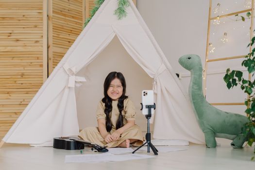 Smiling girl in a tent playing ukulele with smartphone setup, perfect indoor playtime.