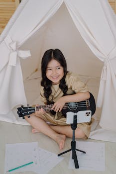 Young girl playing ukulele inside a teepee tent while recording a video on her smartphone.