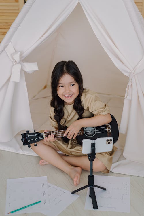 Chica joven con vestido beige sonriendo y sosteniendo un ukelele mientras graba TikTok.