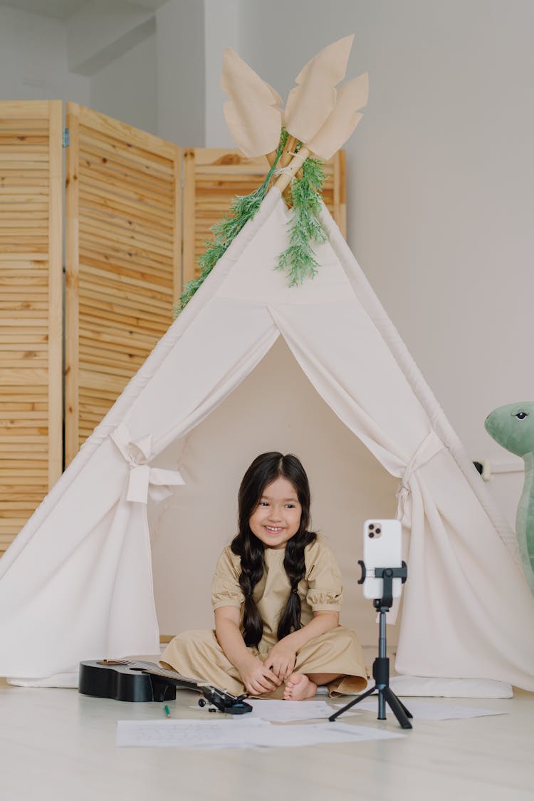 A Cute Girl In Beige Dress Smiling While Looking At The Tripod With Mobile Phone