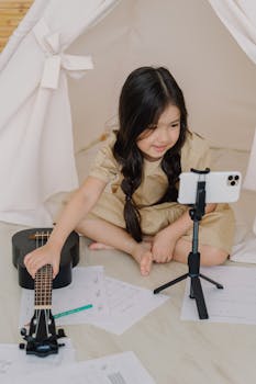 Young girl sitting in a tent, playing a ukulele and recording on her phone.