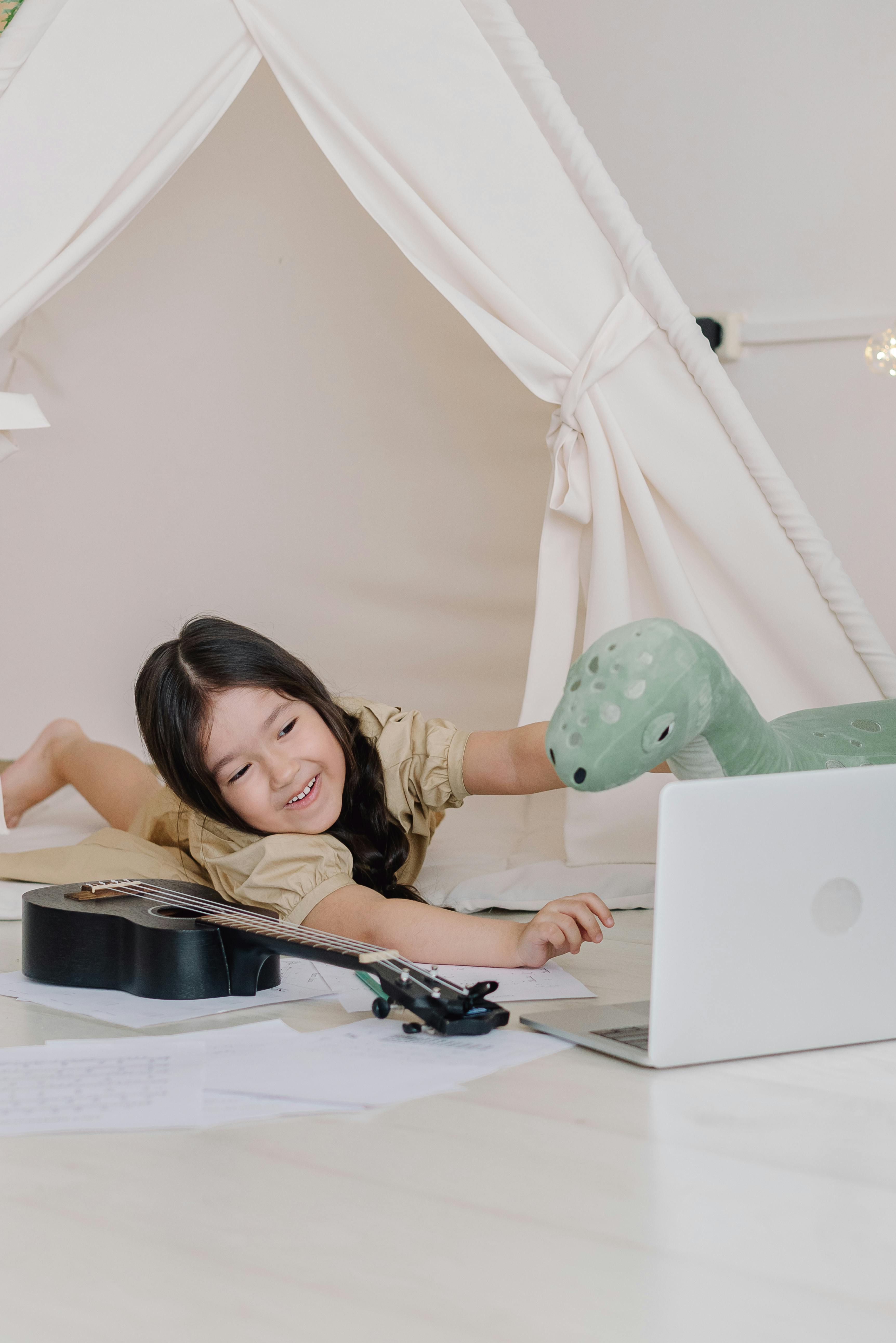 A happy child with a ukulele video chatting in a cozy indoor tent space.