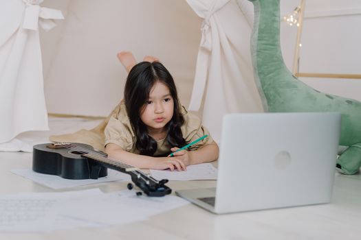 A young girl lies on the floor with a laptop and ukelele, focused on studying indoors.