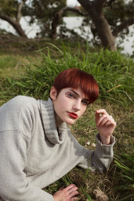 Portrait of a young woman with red hair, in a gray sweater, lying on grass with trees in the background.