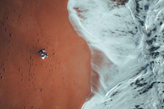 Picturesque drone view of unrecognizable travelers standing on sandy shore washed by foamy waves and admiring stormy ocean