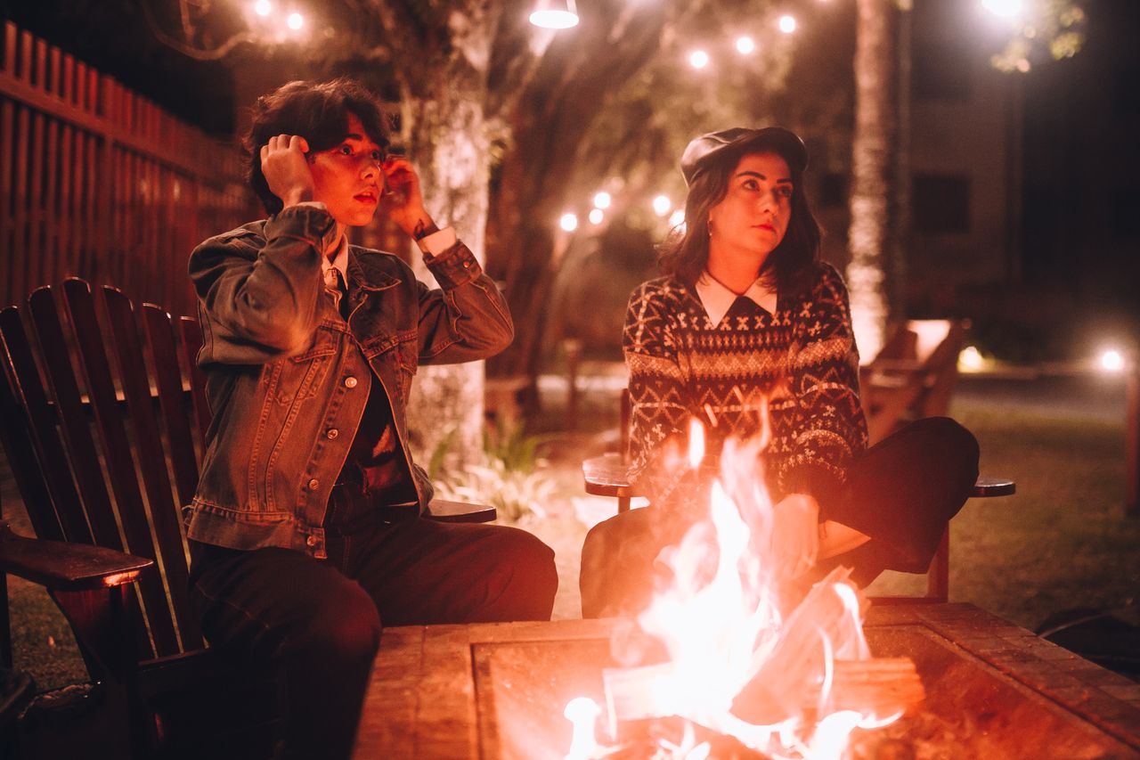 Two friends sitting in Adirondack chairs around a square fire pit table with warm string lights above a fenced backyard at night
