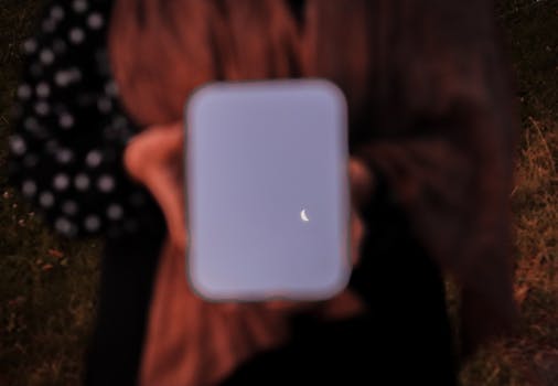 Blurred female holding a mirror reflecting a crescent moon during twilight outdoors.