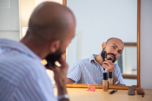 A man in a striped shirt grooms his beard while looking in the mirror indoors.