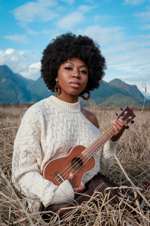 Free A Woman Wearing Knitted Sweater Using a Ukulele While Sitting on the Dry Hay Field Stock Photo