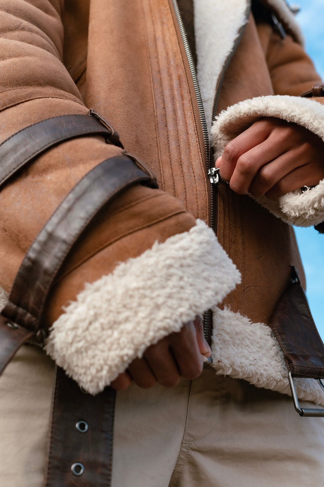 Close-up of hands zipping a tan shearling-lined suede jacket with leather strap details
