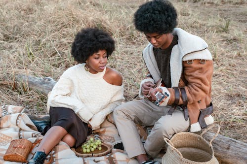 A Woman in White Sweater Looking at the Man Playing Ukulele while Sitting on Grass Field