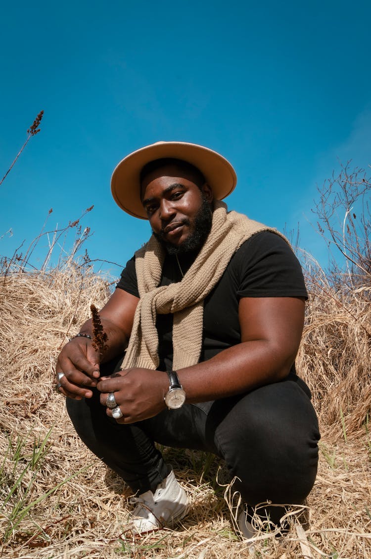 A Man In Black Shirt And Pants Crouching On The Field With Dried Grass