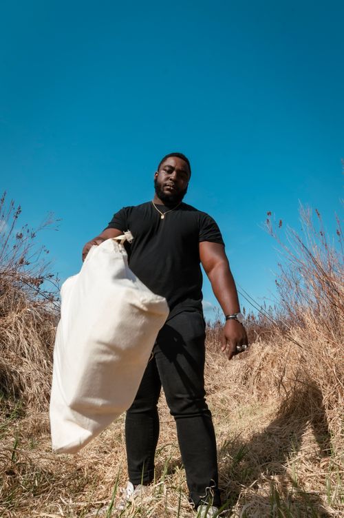 Free A man carrying a tote bag stands in a sunlit grassy field under a clear blue sky. Stock Photo