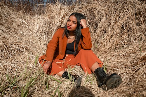 Young Woman in Orange Coat and Pants Sitting on Dry Grass 