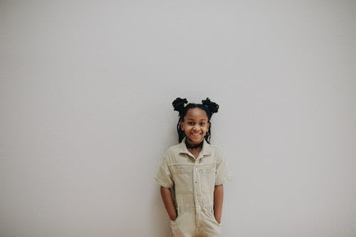 Smiling Girl Wearing Beige Jumper Overall with Pigtails Standing Near a Wall