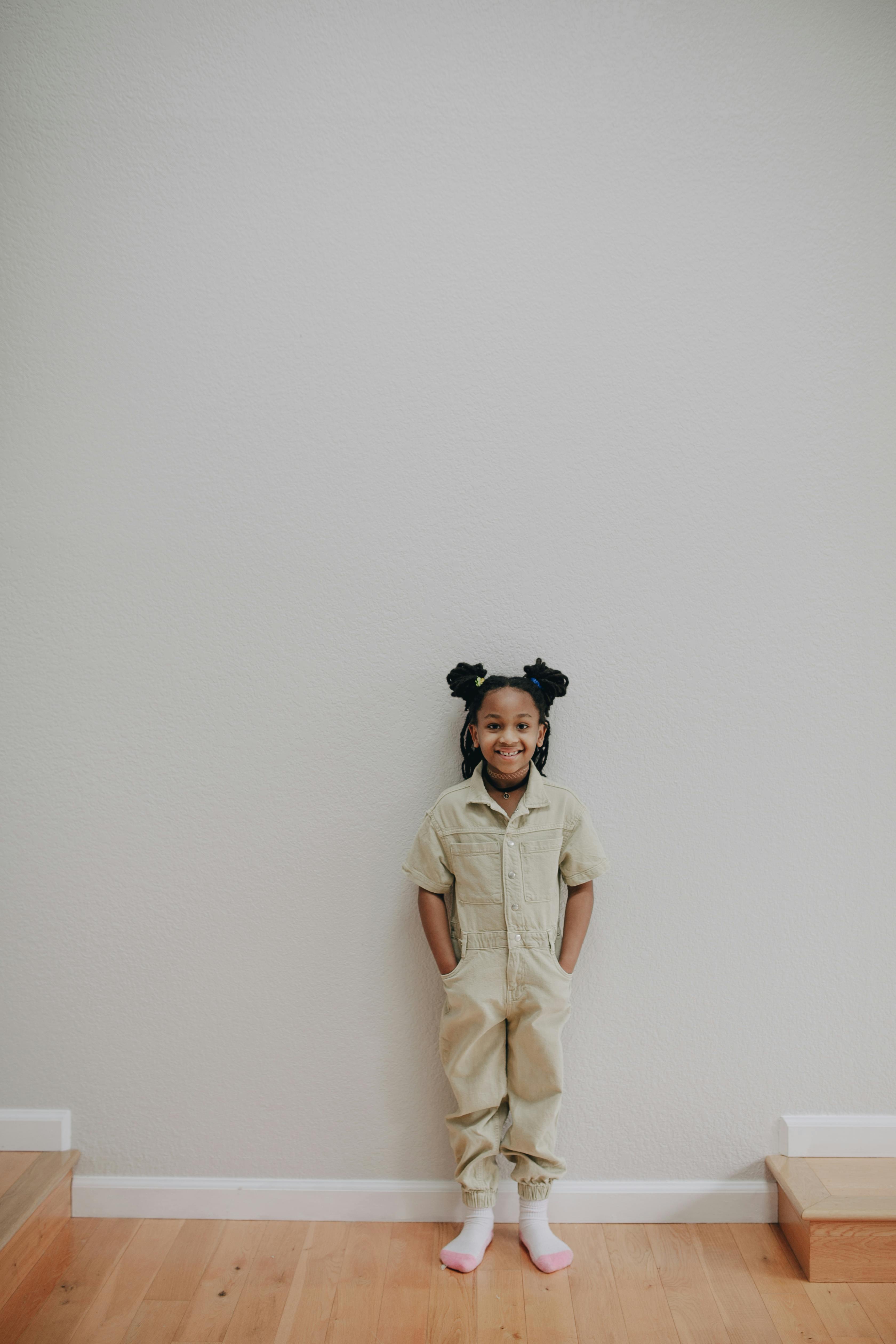 child in beige overall standing on wooden flooring beside a wall
