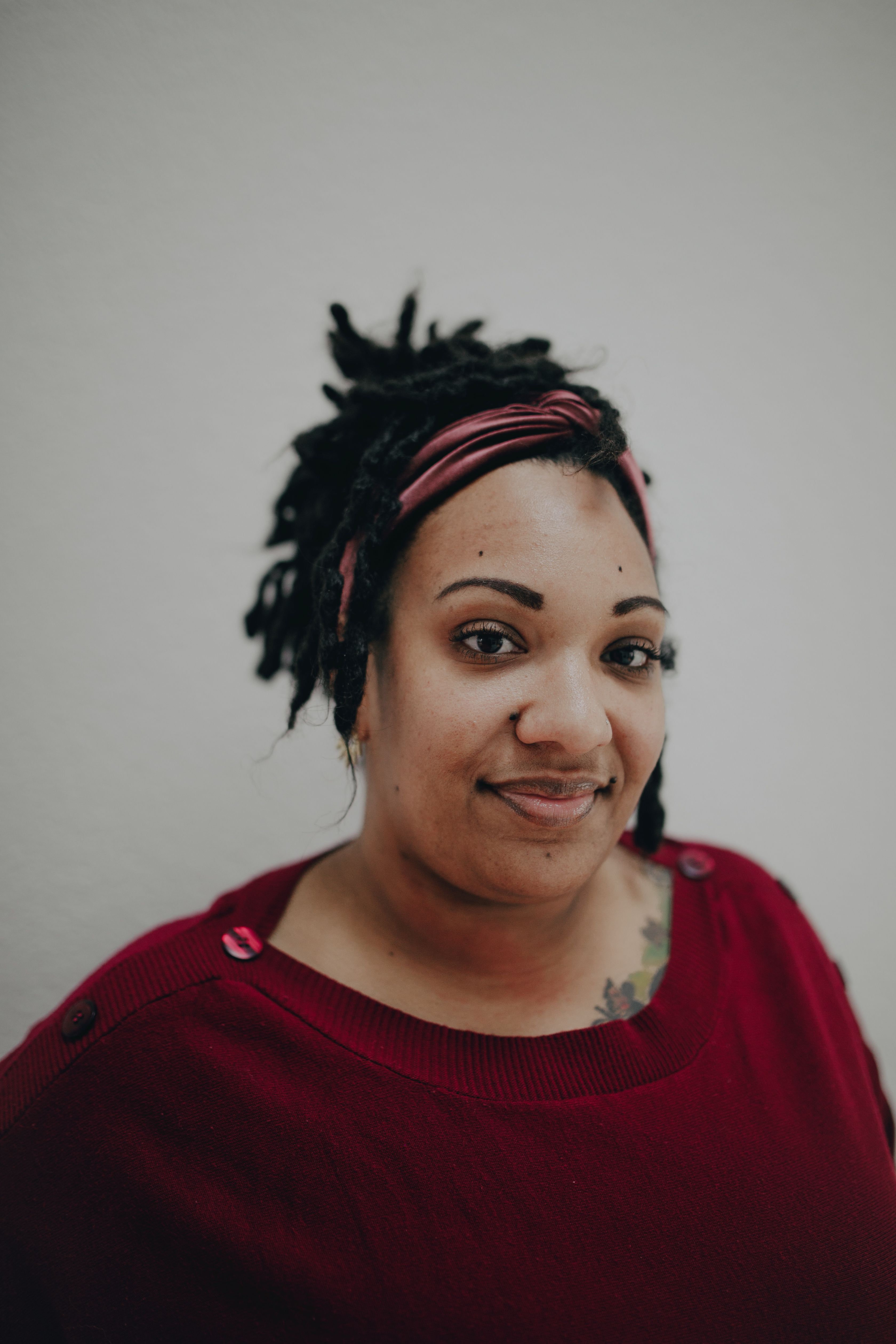 Portrait of a smiling woman wearing a red blouse with dreadlocks and a headband against a light background.
