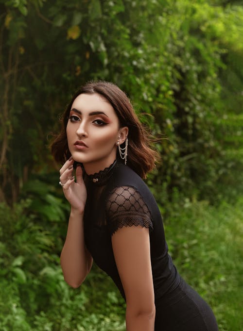 Free Side view of young dreamy woman wearing black elegant blouse with makeup and accessories standing near green bushes and plants in nature while looking at camera in summer day Stock Photo