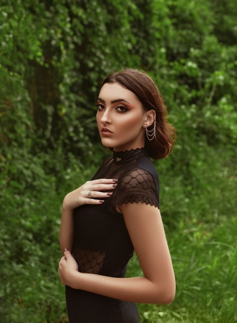 Side view of confident young female wearing black elegant blouse with makeup and accessories standing in park near green shrubs and plants while looking at camera in summer day