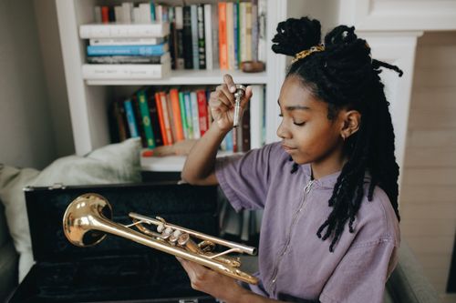 Free A young girl with dreadlocks holds a trumpet indoors, surrounded by books. Stock Photo