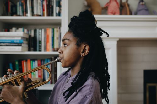 Free A young black girl playing the trumpet indoors, surrounded by books. Creative and inspiring scene. Stock Photo