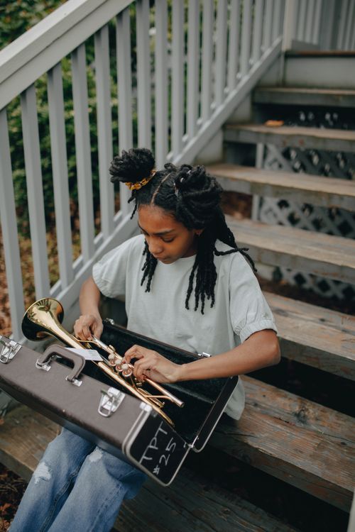 Free African American girl sitting on stairs, holding a trumpet from its case outdoors. Stock Photo