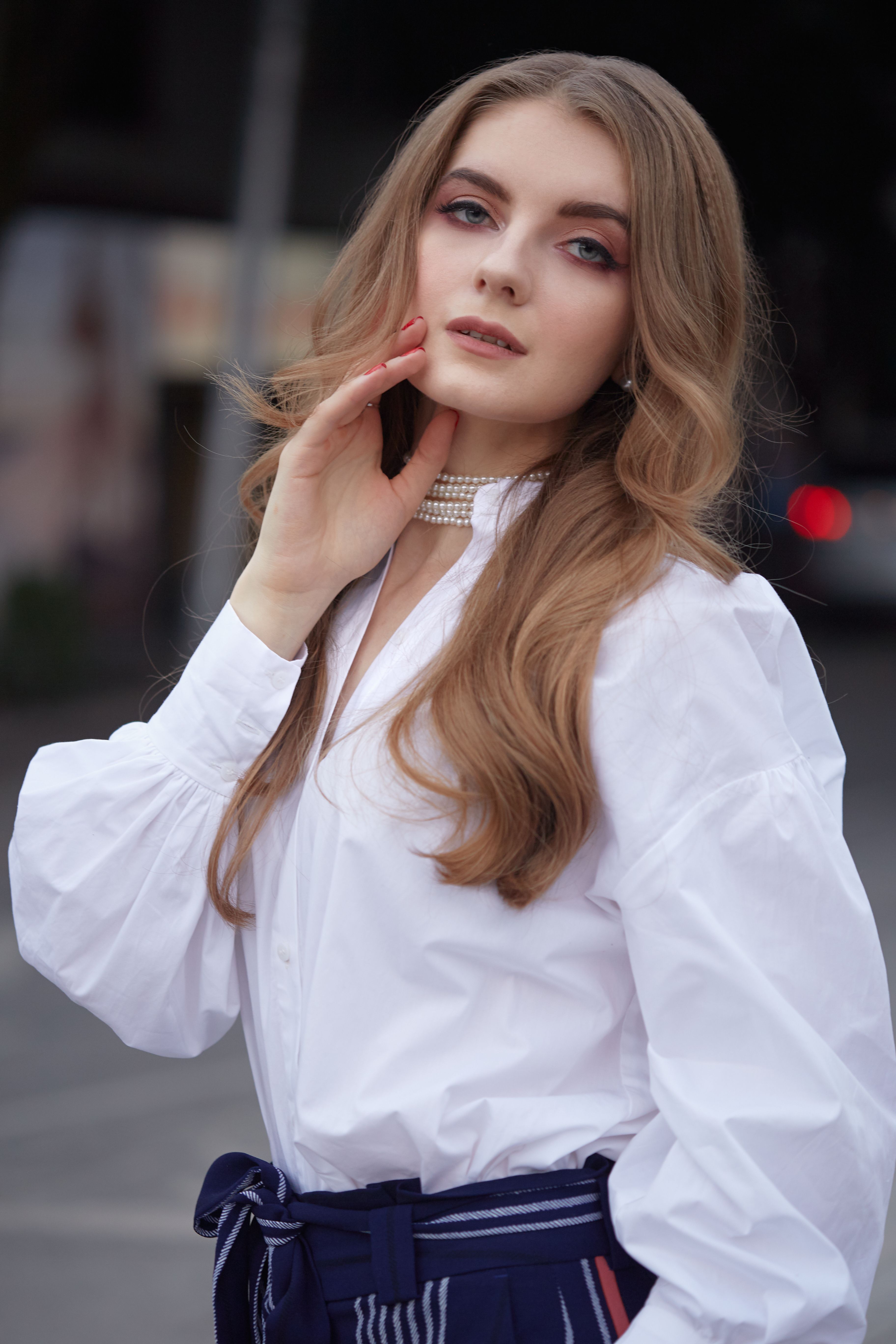 Sensual female with long blond hair and makeup in white blouse standing on city street and looking at camera