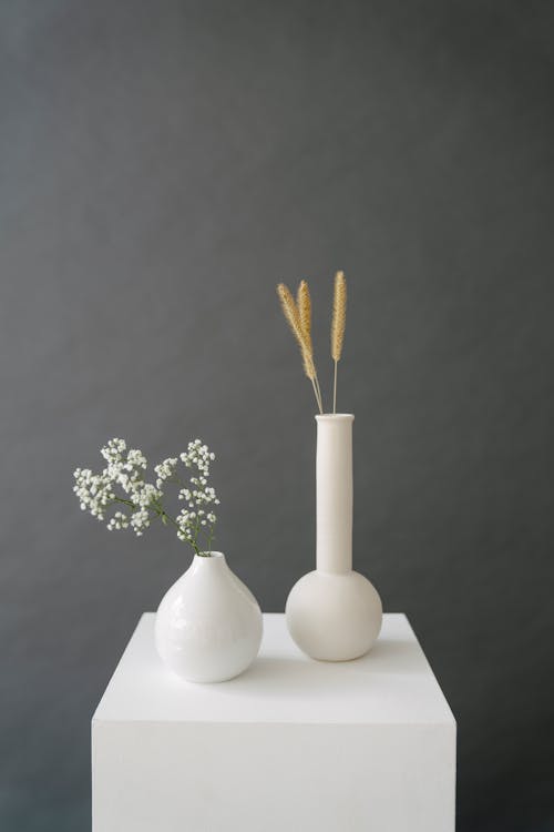 Ceramic vases with wheat ears and blooming branch placed on pedestal in studio against gray background