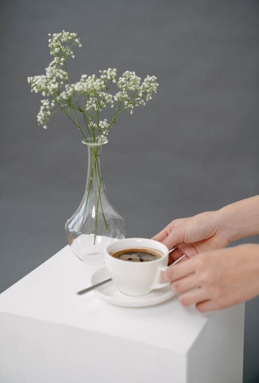 Free High angle of crop anonymous female taking cup of aromatic Americano placed near vase of blooming branches on white cube Stock Photo
