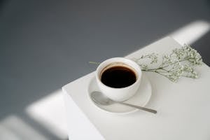 From above of ceramic cup of hot Americano coffee placed on saucer near spoon and twig of plant on white cube