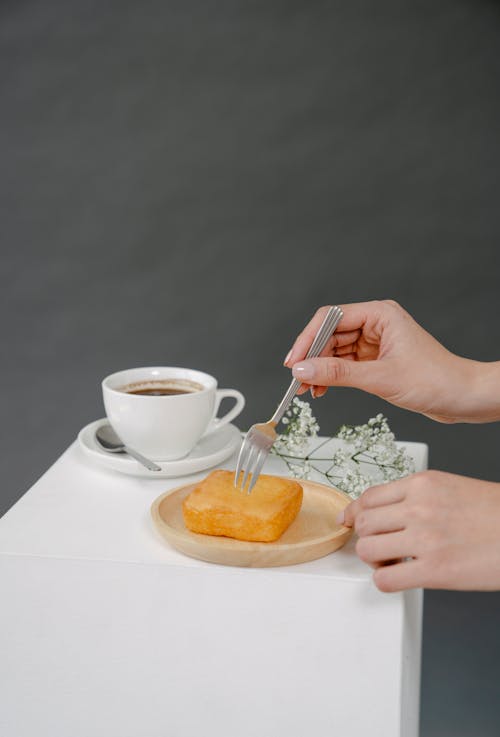 Crop anonymous female eating sweet sponge cake served on wooden plate near cup of coffee on gray background in studio