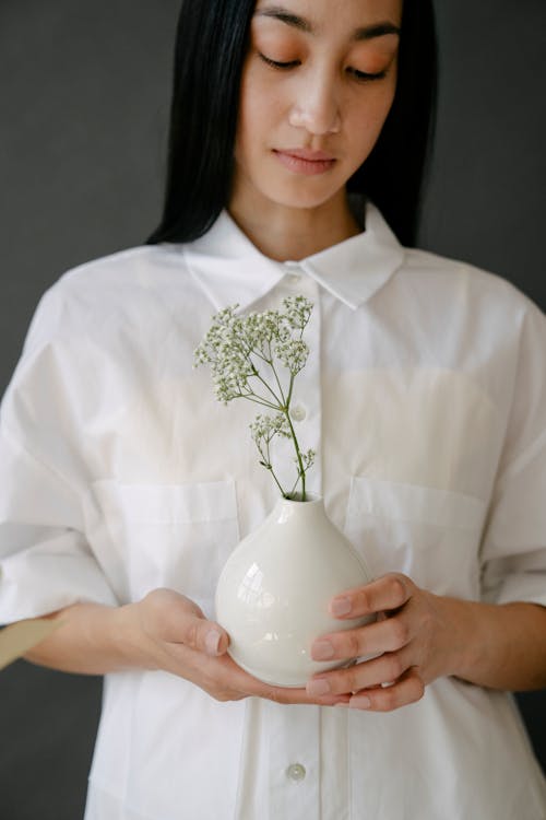 Crop Asian female in white clothes standing on gray background with thin twigs in ceramic vase in light modern studio