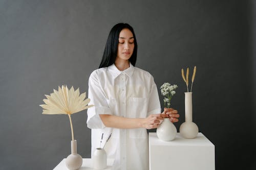 Attractive Asian female in white clothes arranging twigs in vase in light studio with decorative dried plants on gray background