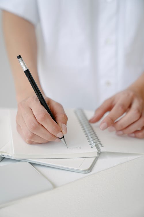Crop unrecognizable female in white wear taking notes in notepad with black pen while sitting at table in light room