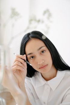 Serious ethnic woman in white clothes looking at camera and holding pen near blurred dried twig in transparent vase