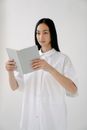 Asian female in white shirt opening notebook and looking away while standing on white background