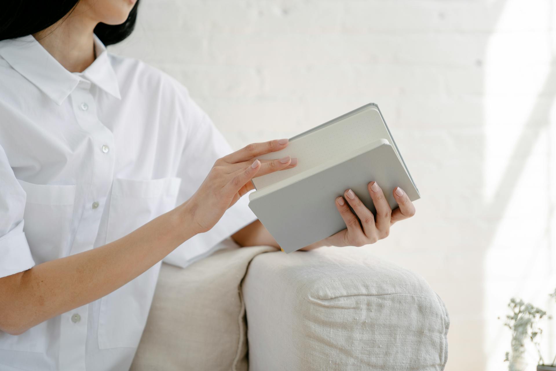 Crop unrecognizable female in white shirt sitting on sofa and opening notepad in light room